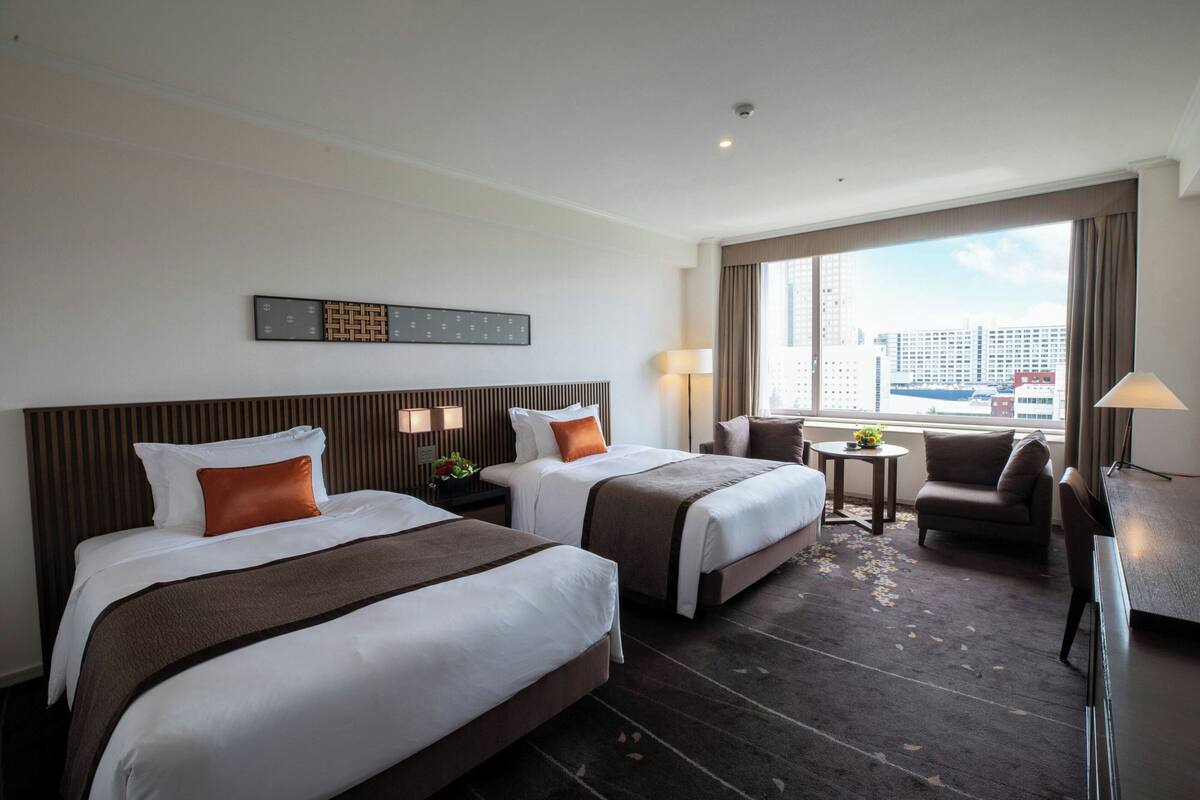 A hotel room with two beds, a desk, a chair, and a view of the city.
