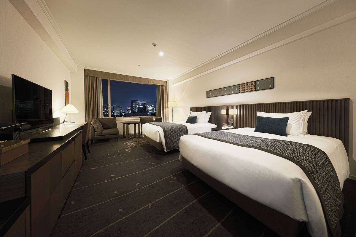 A hotel room with two beds, a desk, a TV, and a view of the city at night.