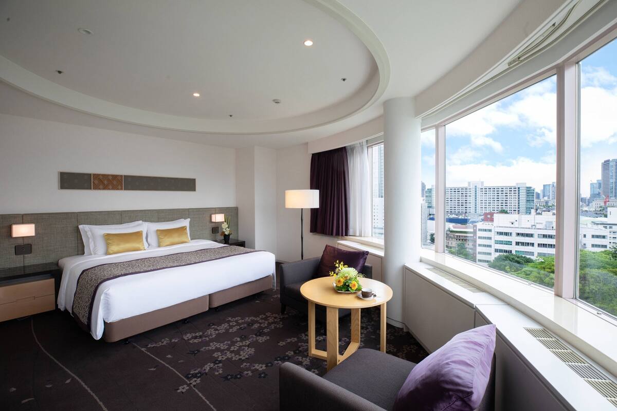 A hotel room with a large bed, a small round table with a flower vase, a sofa, and a view of the city through large windows.