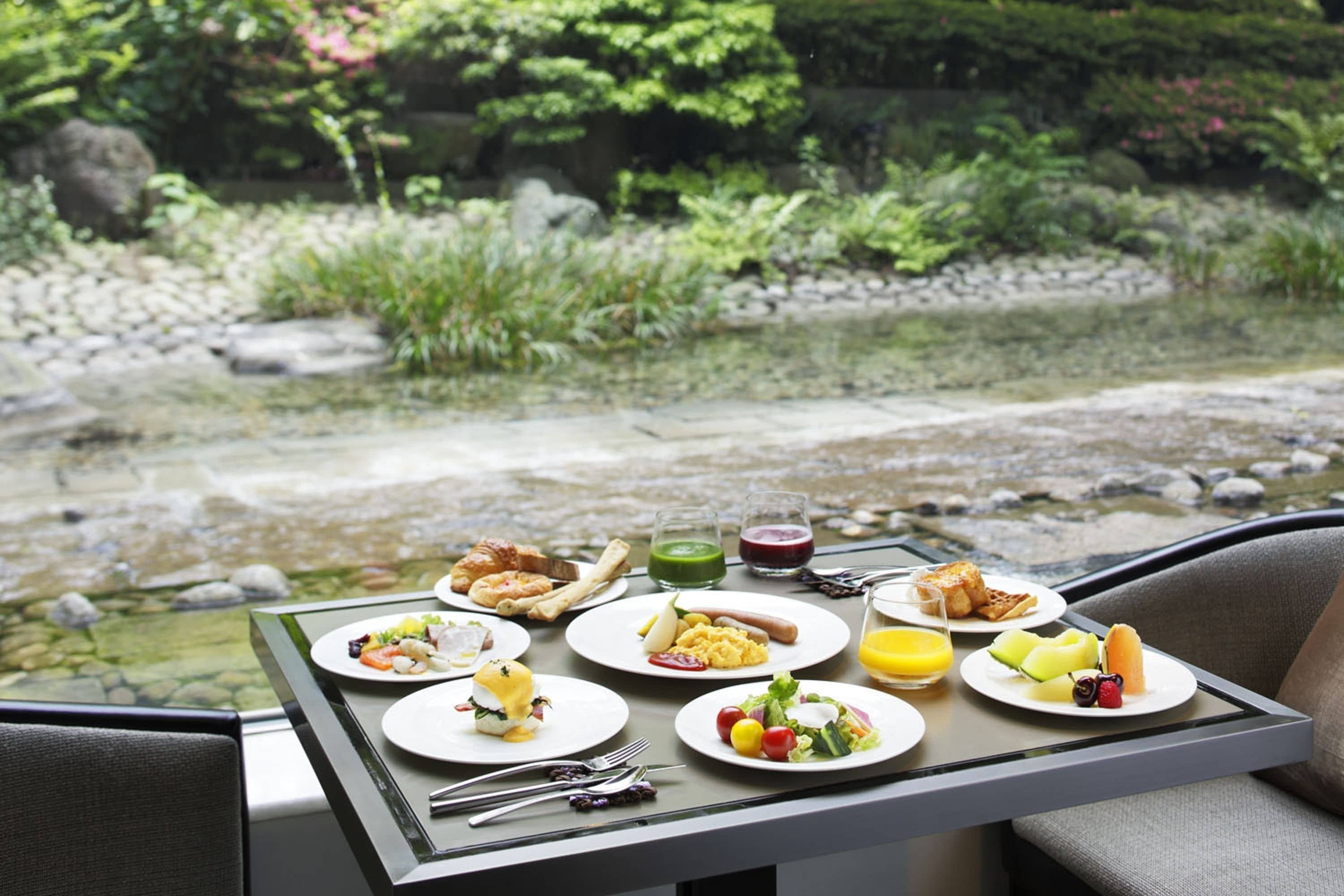 A breakfast table with plates of food and drinks, overlooking a garden with a pond.