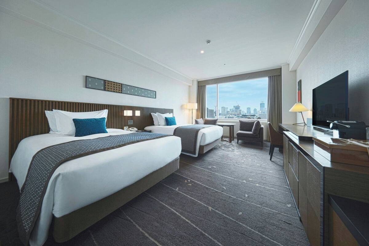 A hotel room with two beds, a desk, a TV, and a view of the city skyline.