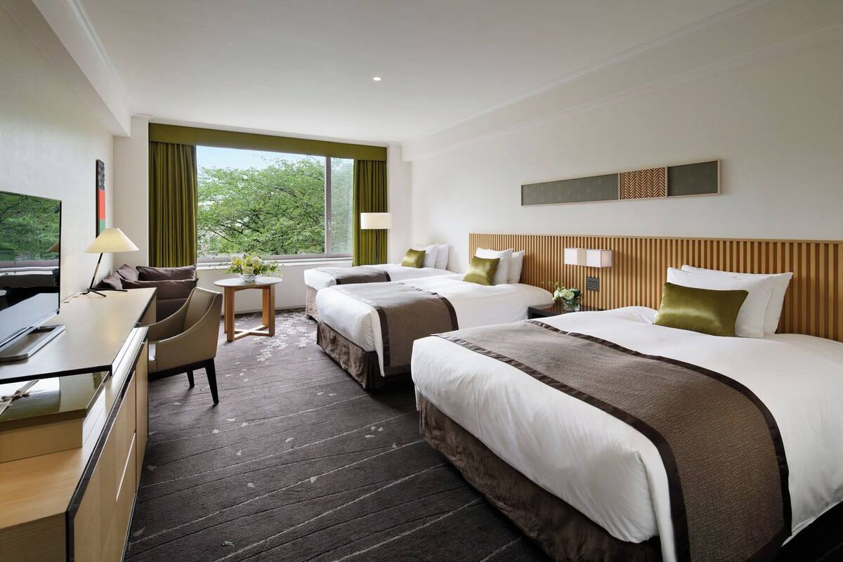 A hotel room with two beds, a desk, a chair, a small table with a vase of flowers, and a view of greenery outside.