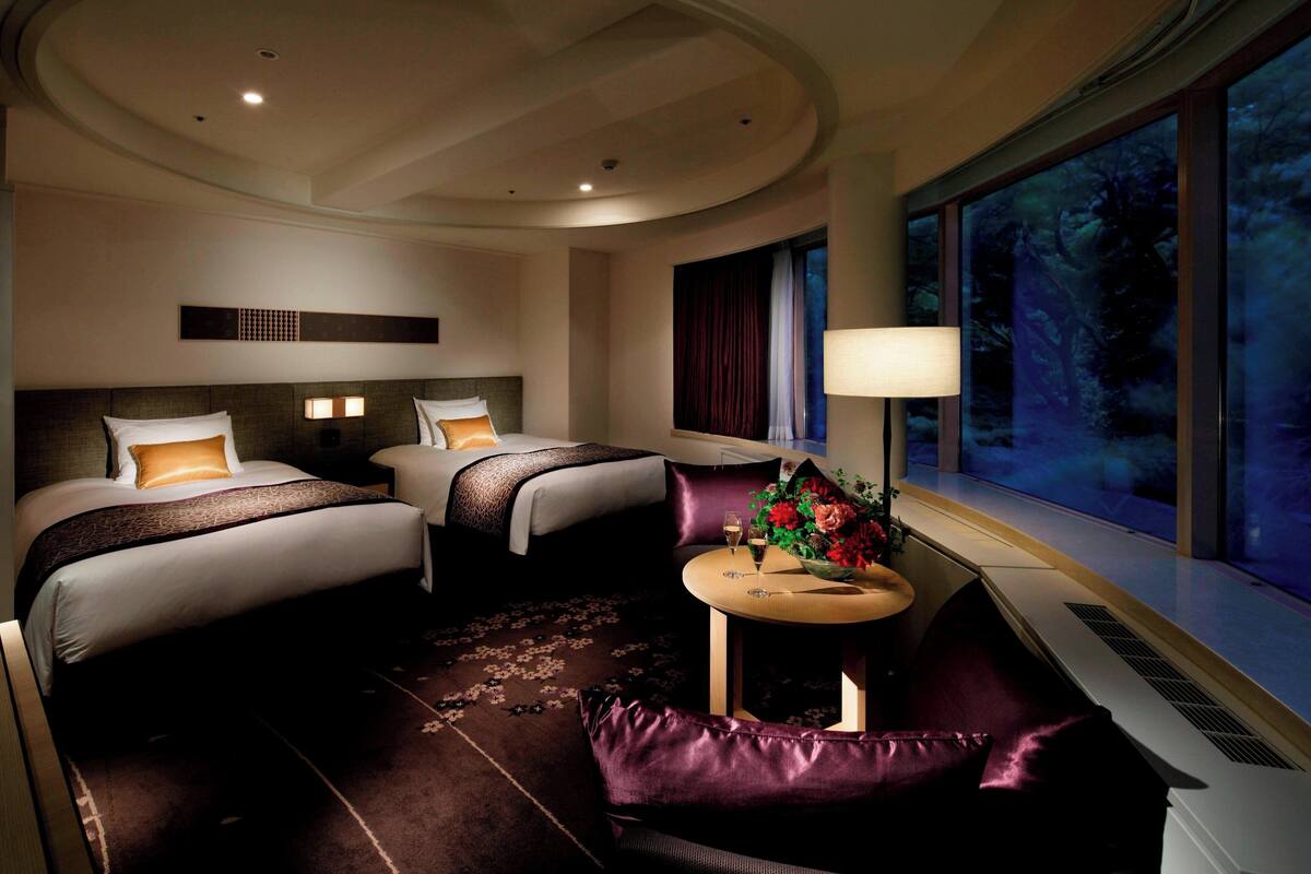 A hotel room with two beds, a sofa, a small table with a vase of flowers, and a large window with a view of trees.