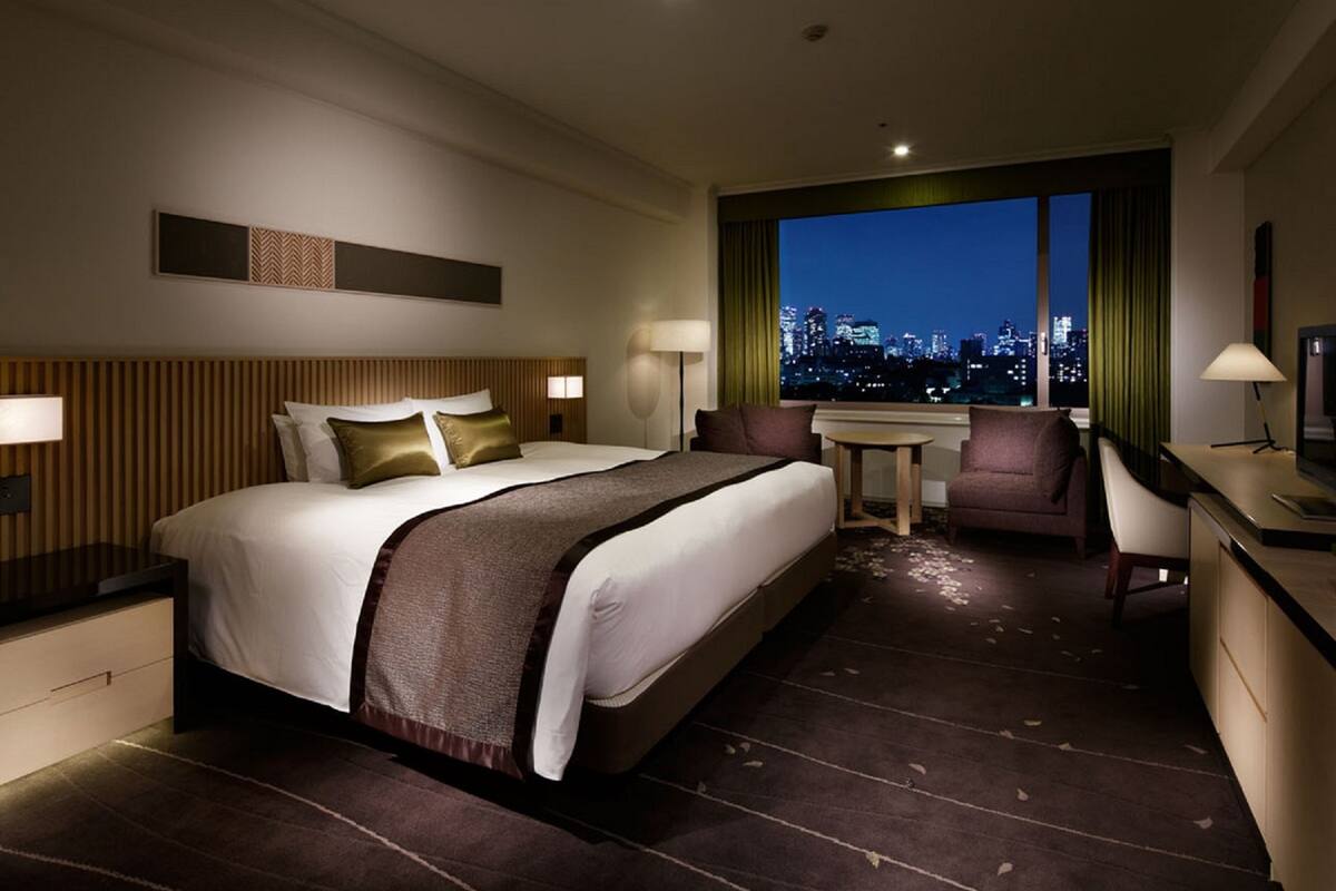A hotel room with a large bed, a desk, a chair, and a view of a cityscape at night.