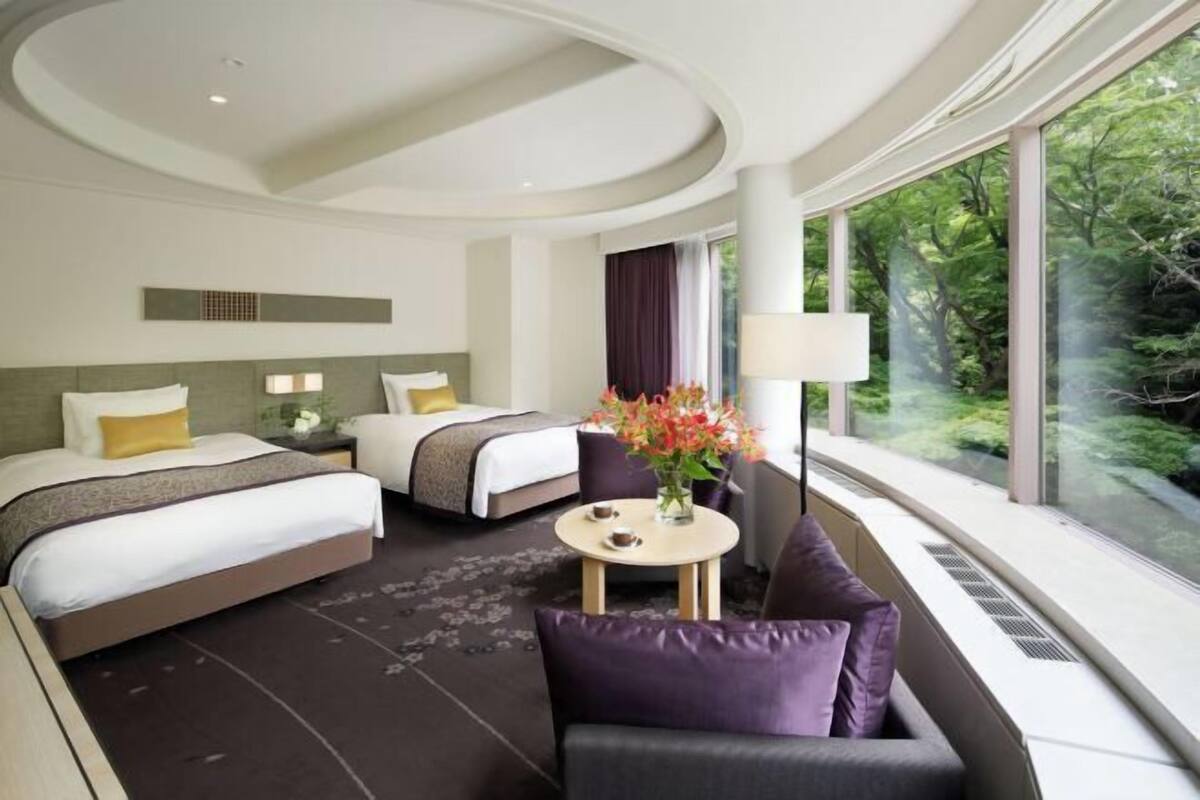 A hotel room with two beds, a sofa, a small table with a vase of flowers, and large windows offering a view of greenery.
