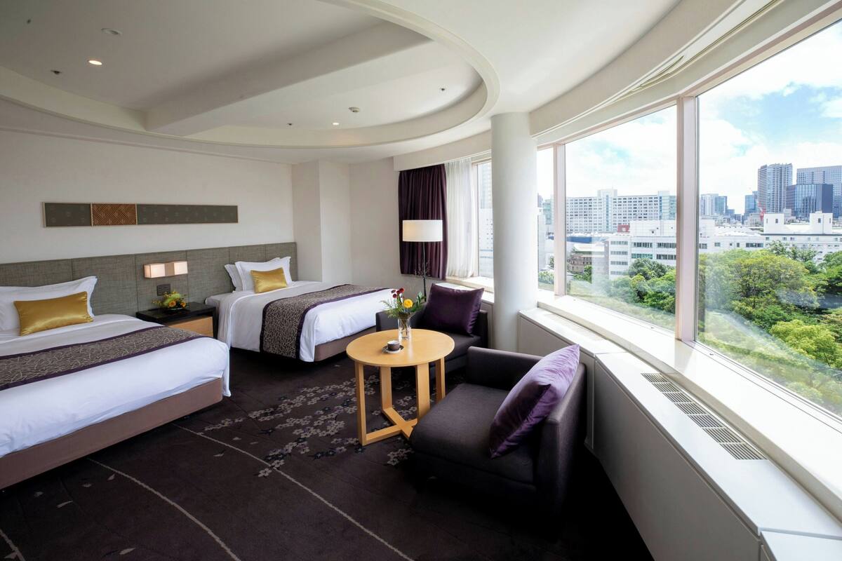 A hotel room with two beds, a small round table, a chair, and a large window with a city view.