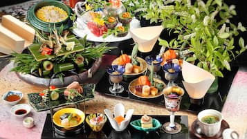 A variety of dishes and drinks on a table, including sushi, sashimi, and other Asian cuisine, with a backdrop of green plants.