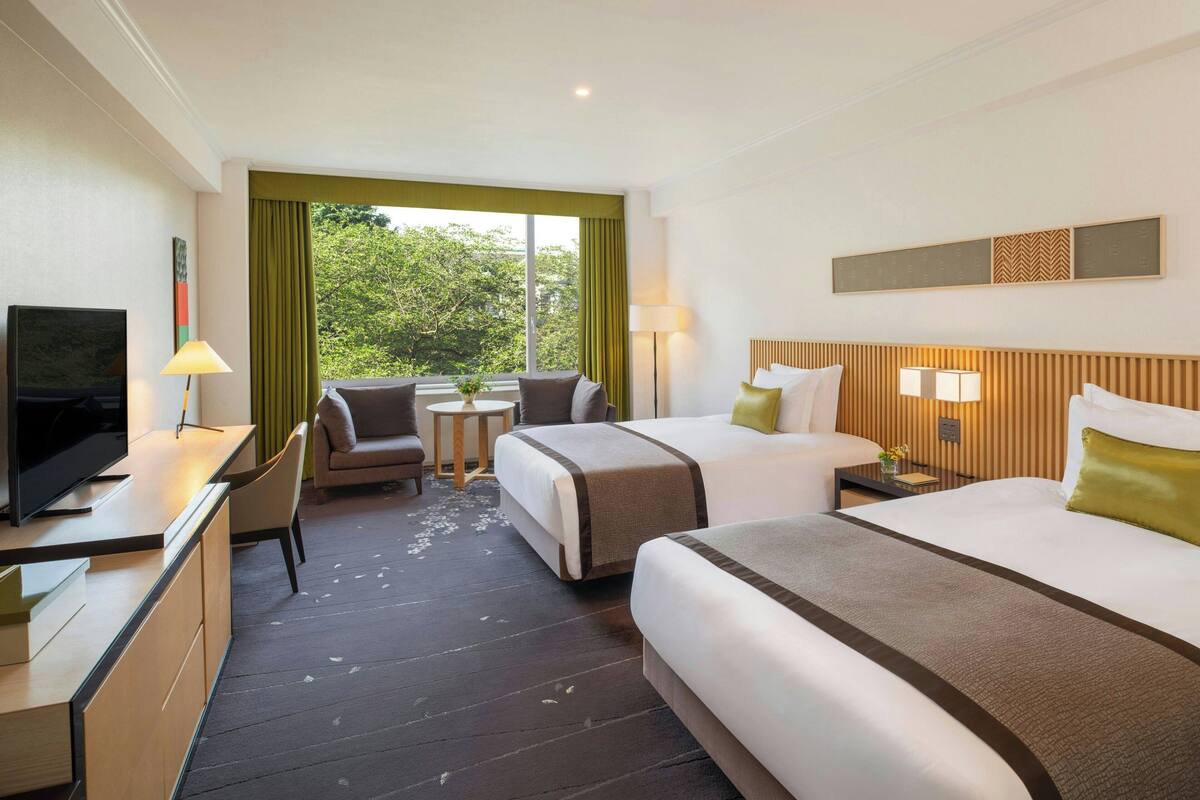 A hotel room with two beds, a desk with a TV, a small table with a lamp, and a view of greenery through the window.