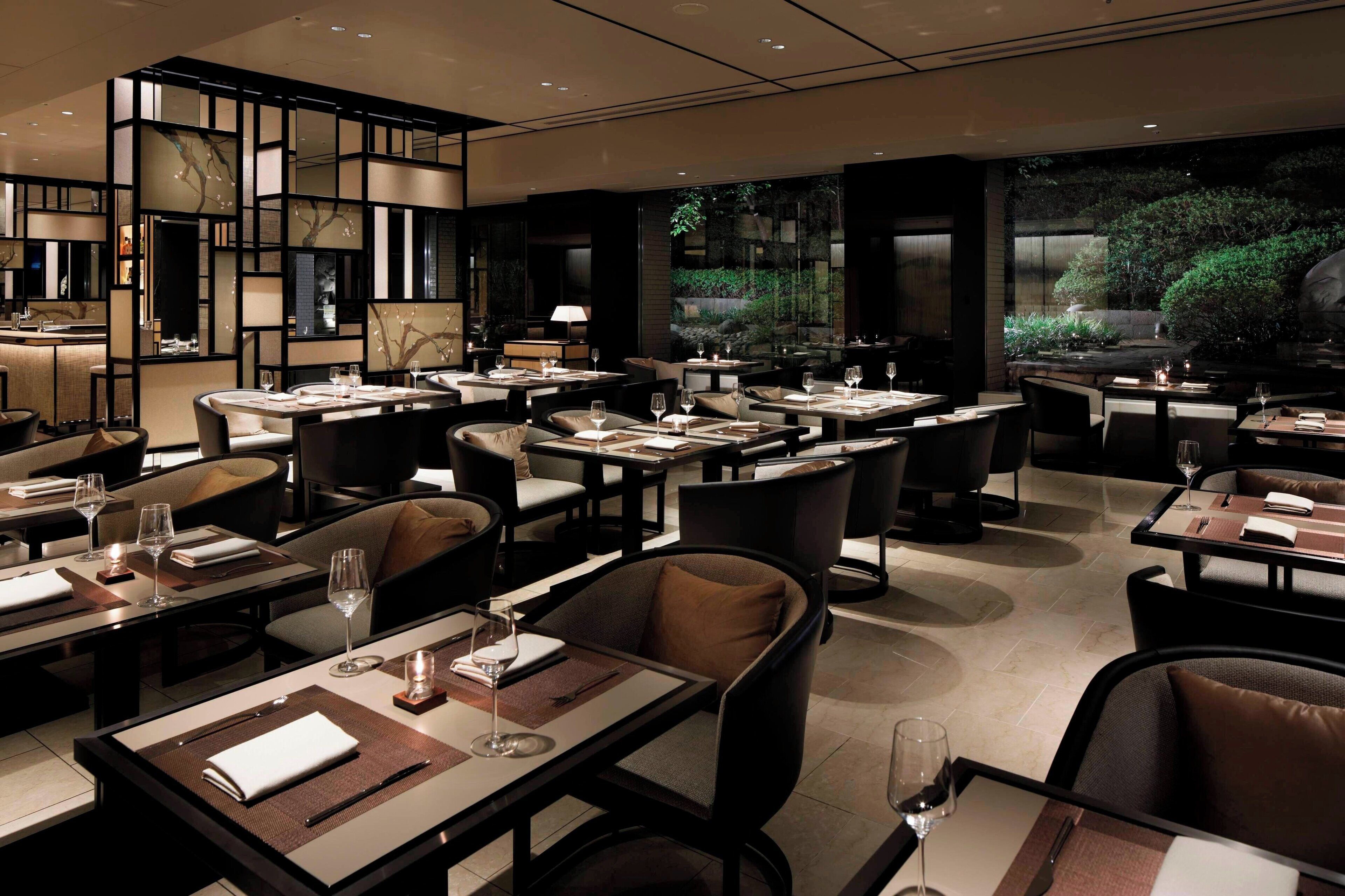 A modern restaurant with tables set for dining, featuring a minimalist design and large windows overlooking a garden.