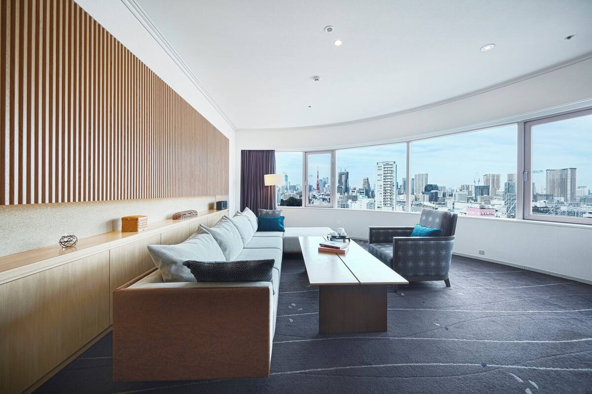 A modern hotel room with a curved ceiling, a sofa set, a dining table, and a view of the city skyline through large windows.