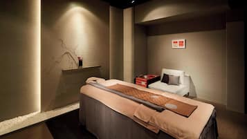 Ein Spa-Raum mit einer Massageliege, einem kleinen Regal mit Dekoration und einem weißen Stuhl.