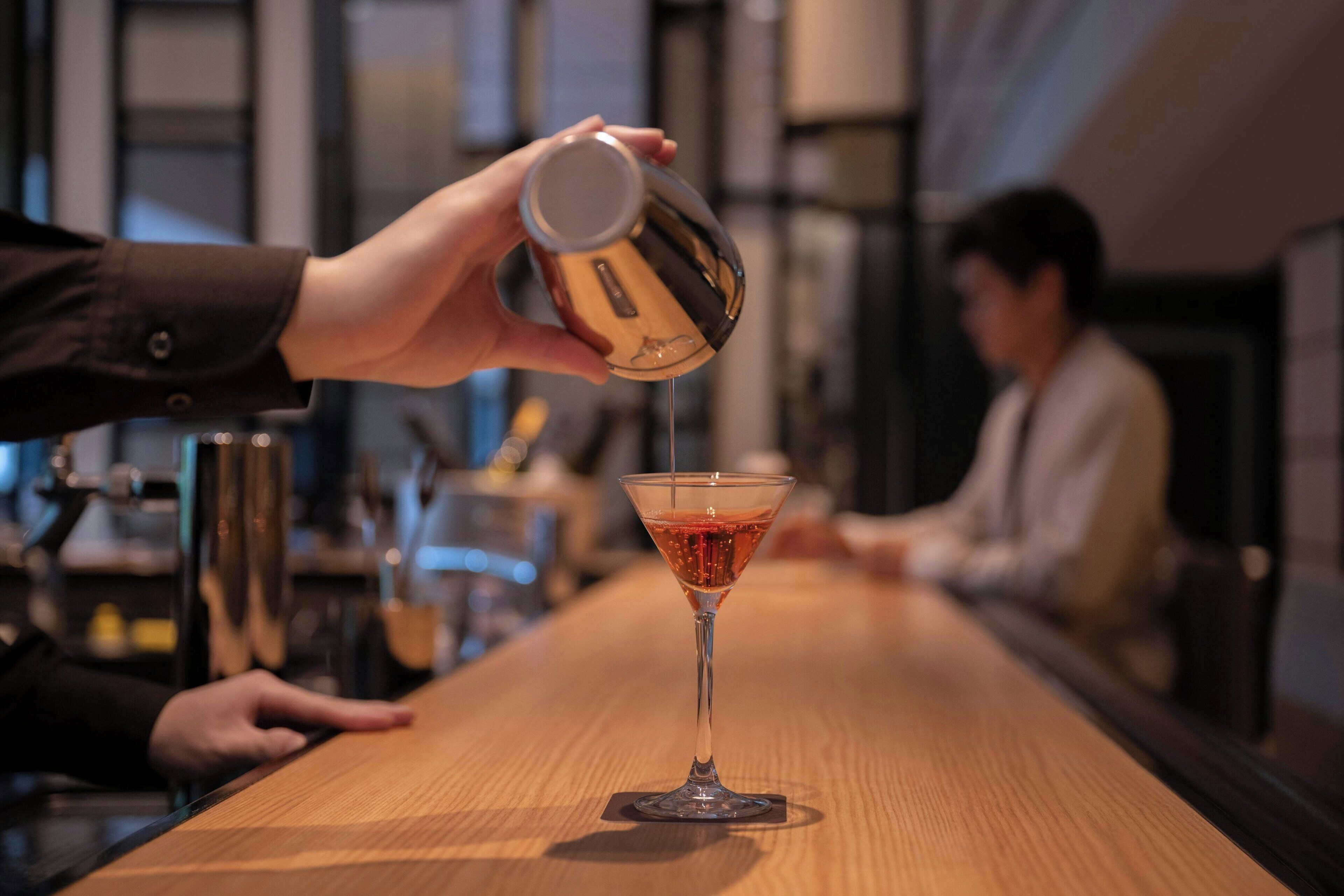 En hand häller upp en drink i ett martiniglas på en bardisk av trä.