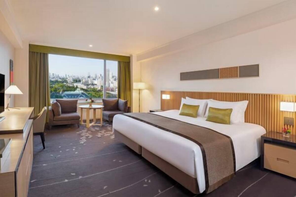 A hotel room with a large bed, a desk, a chair, a small table, and a view of the city.