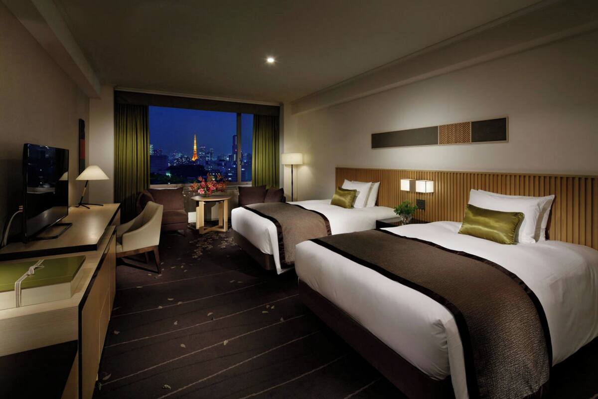 A hotel room with two beds, a desk, a chair, a TV, and a view of a cityscape at night.