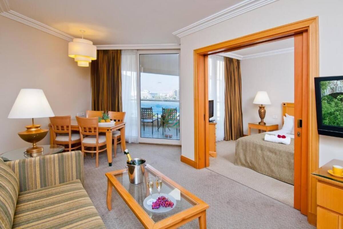 A hotel room with a sofa, a dining table with chairs, a balcony with a table and chairs, a bed with a TV, and a minibar.