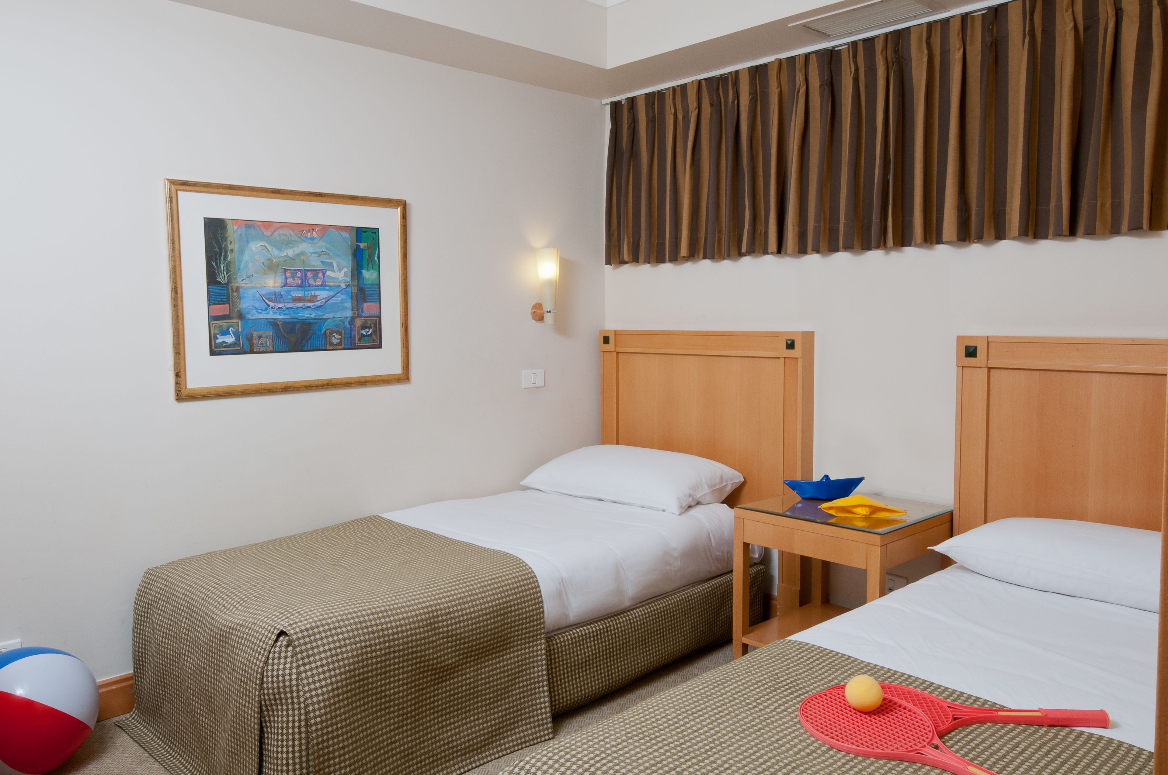 Habitación de hotel con dos camas, un cuadro en la pared, una mesita de noche con un cuenco y un libro, y una pelota de playa en el suelo.