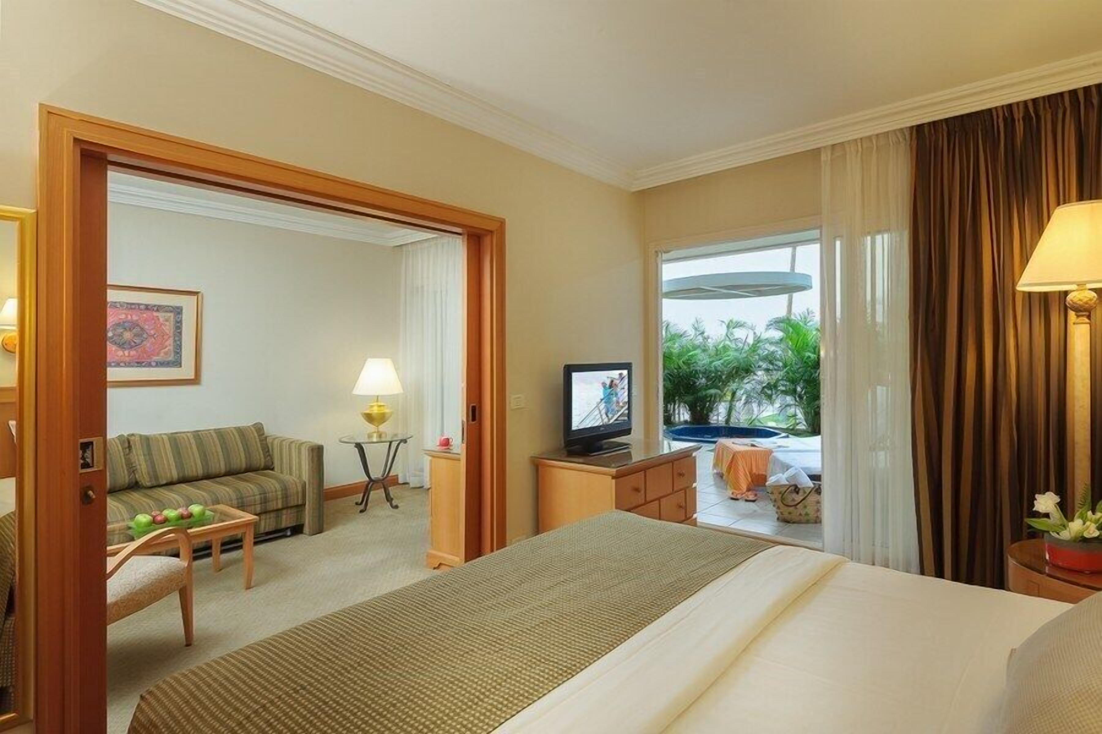 A hotel room with a bed, a sofa, a TV, a small table with a lamp, and a view of the outdoors.
