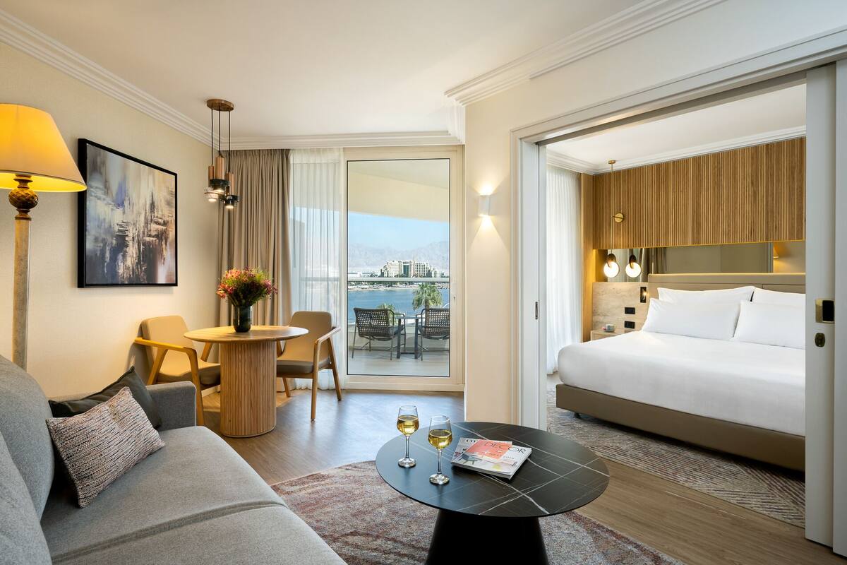 A hotel room with a bed, a sofa, a small table with drinks, a chair, a view of the city, and a balcony with a table and chairs.
