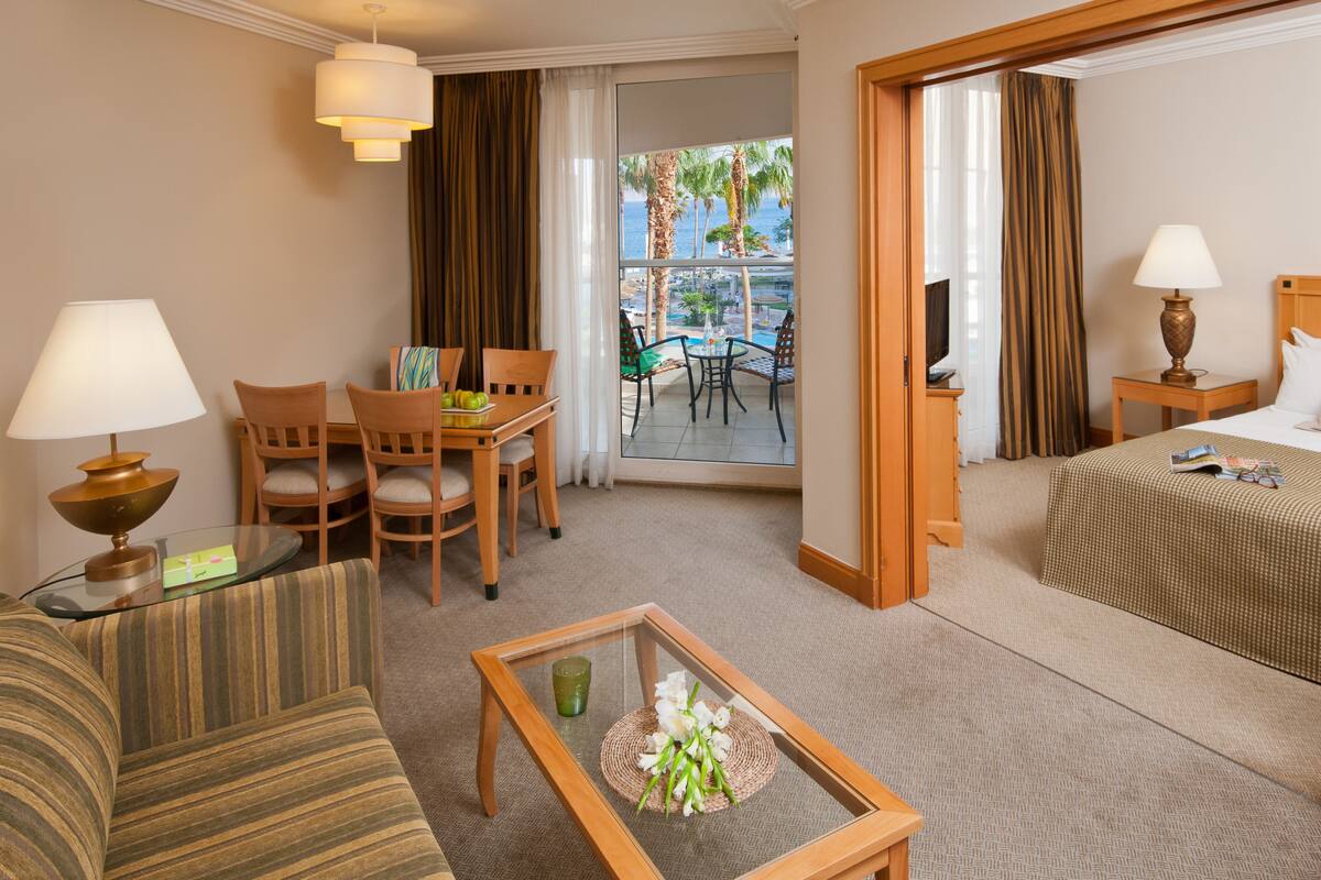 A hotel room with a bed, a sitting area with a table and chairs, a TV, and a balcony with a view of palm trees.