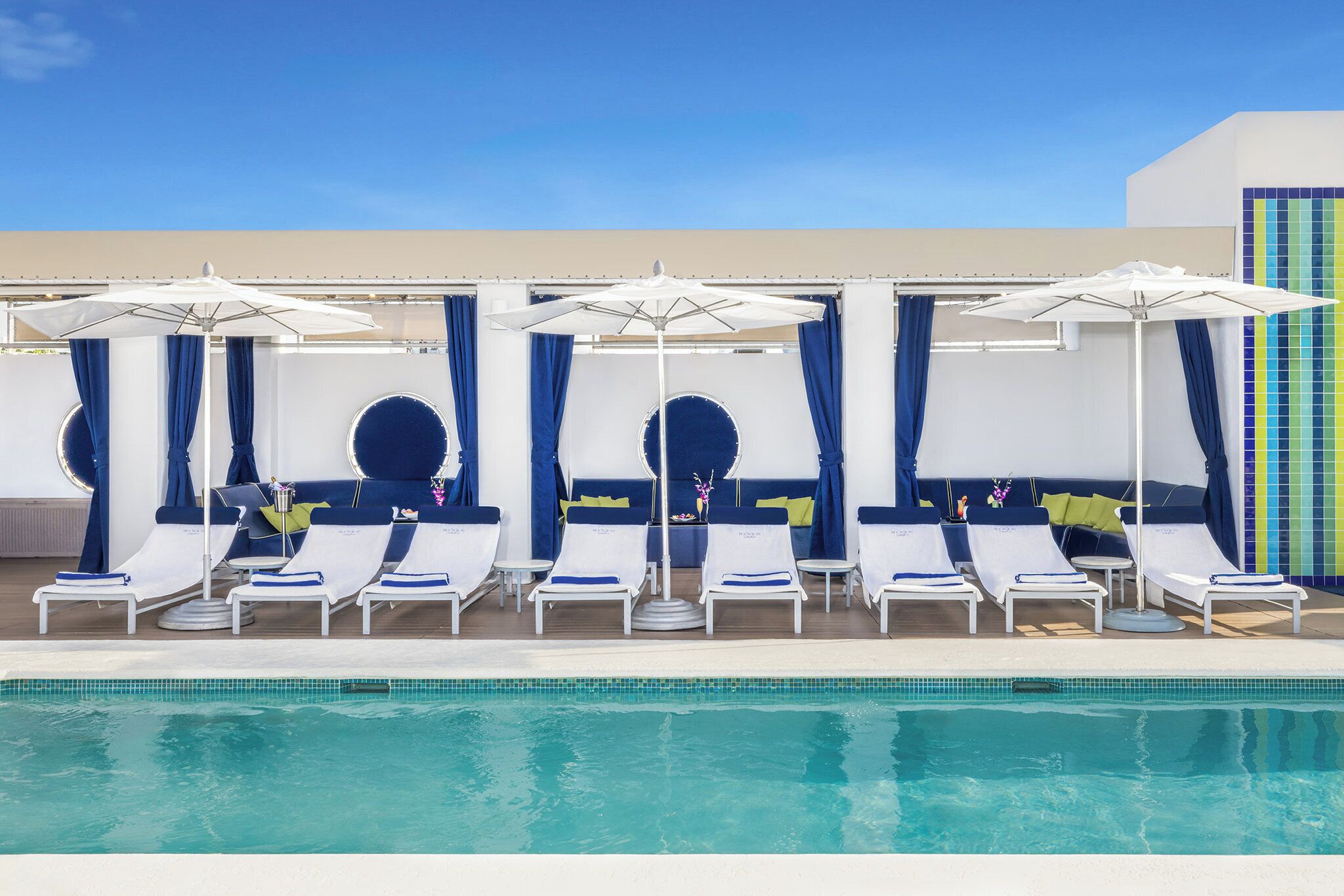 A poolside area with lounge chairs and umbrellas, a clear blue sky, and a tiled wall.