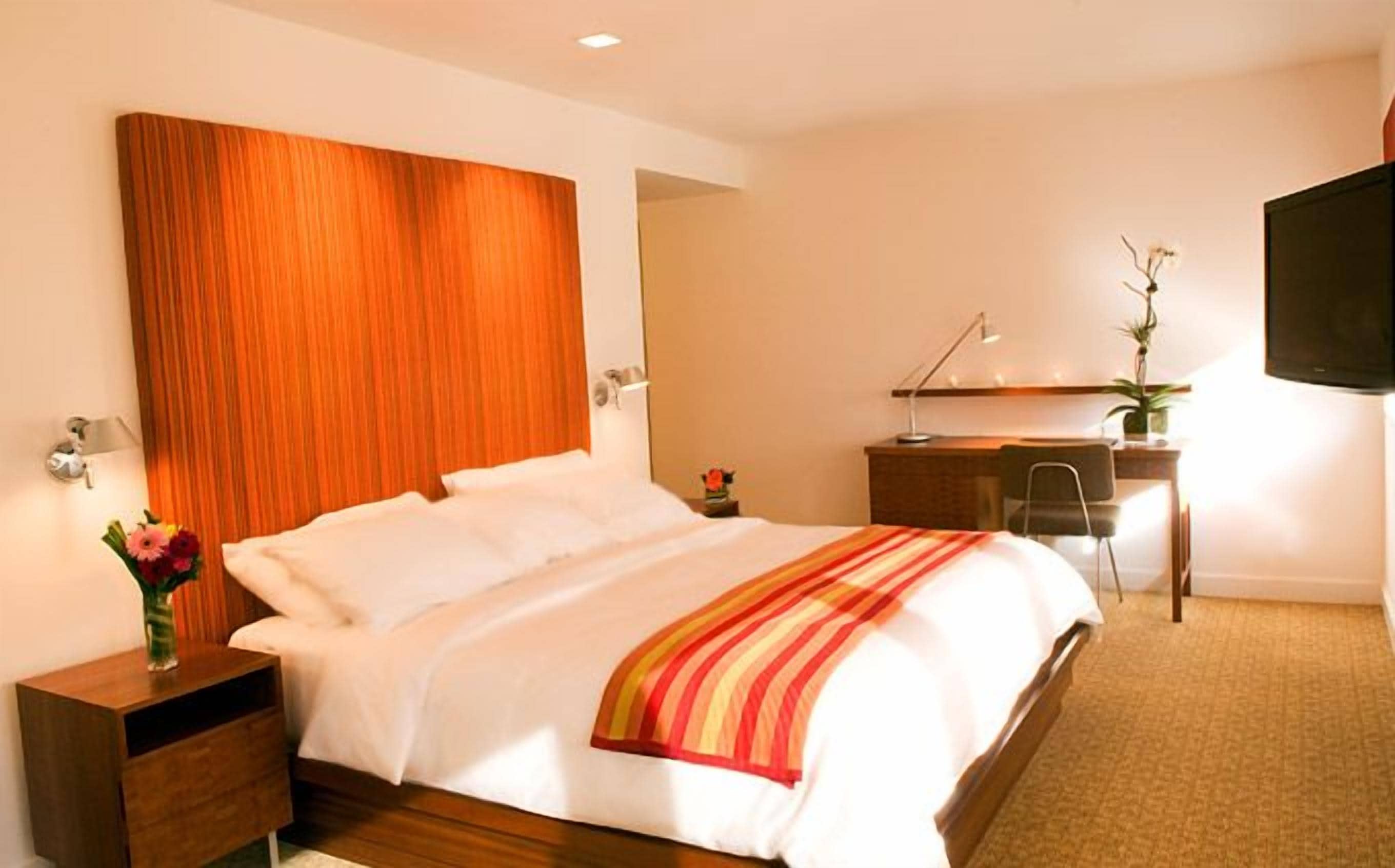A hotel room with a bed, bedside table, desk, chair, and a TV mounted on the wall.