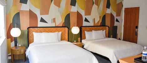 Habitación de hotel con dos camas, cabecero de madera, mesita de noche redonda, lámpara y una pared con arte abstracto.