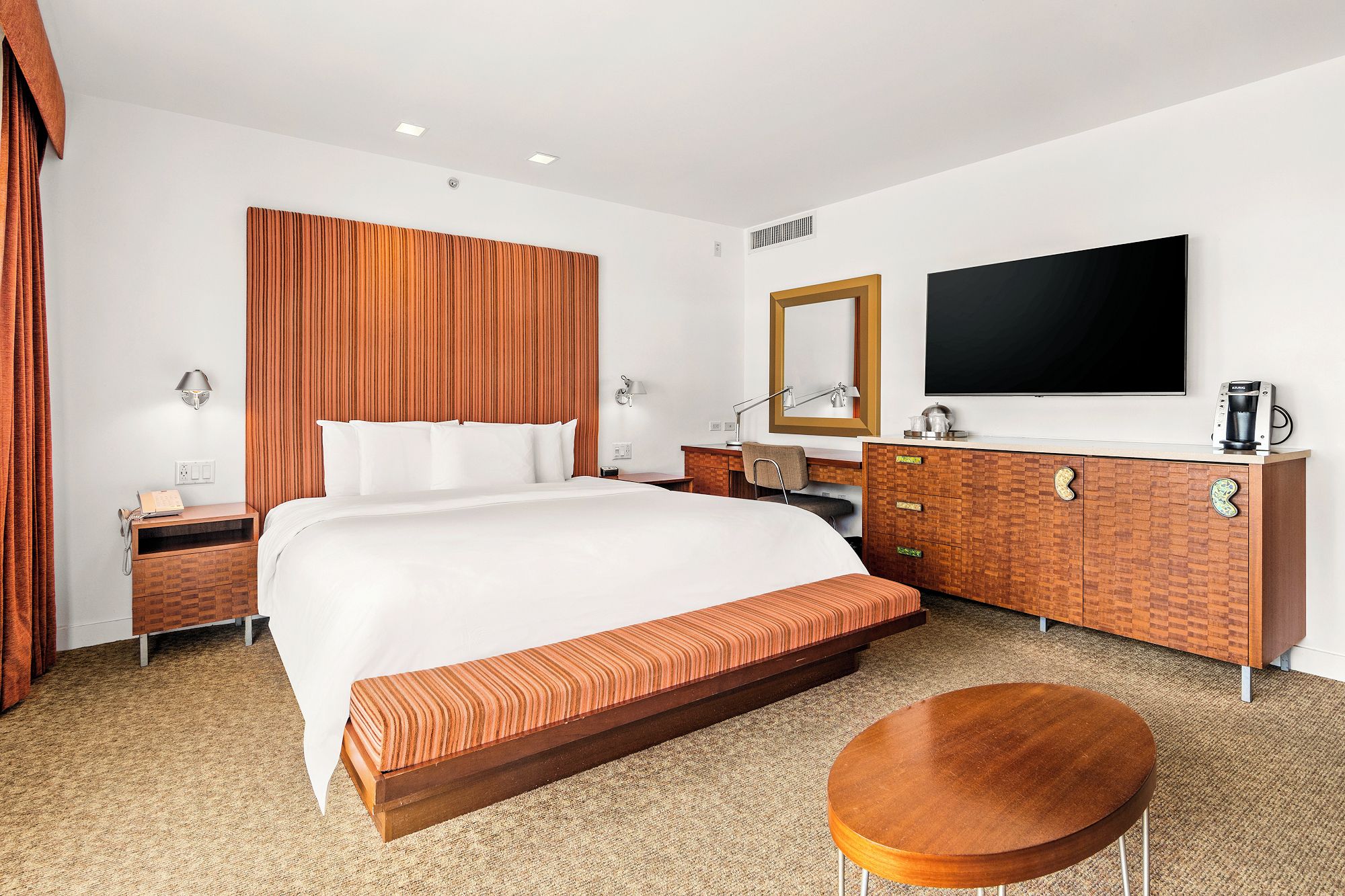 A hotel room with a large bed, a wooden bench, a round table, a TV, a coffee machine, and a desk with drawers.