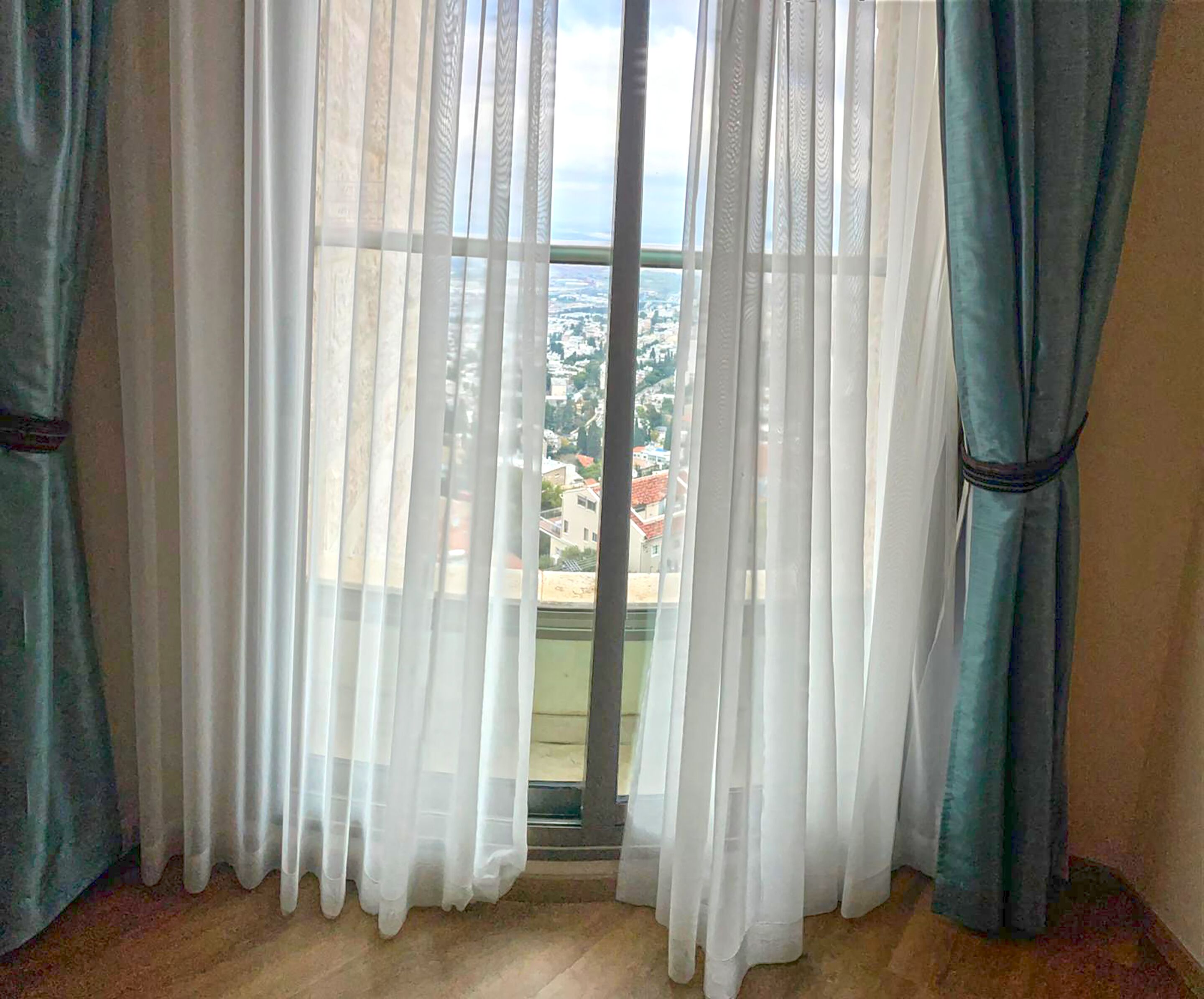 A room with sheer white curtains, a view of a cityscape through a window, and a wooden floor.