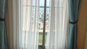 Ein Zimmer mit transparenten weißen Vorhängen, Blick auf eine Stadtkulisse durch ein Fenster und Holzboden.