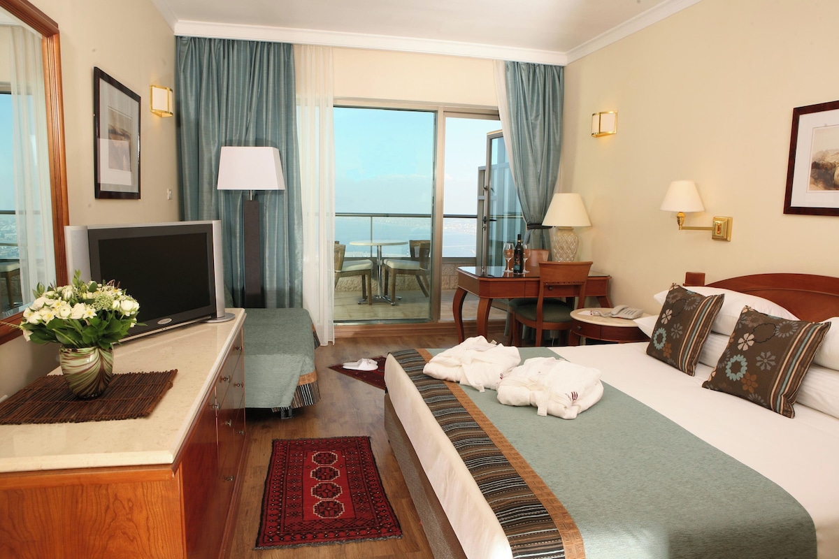 A hotel room with a bed, a desk, a TV, a balcony with a view, and a dining area.