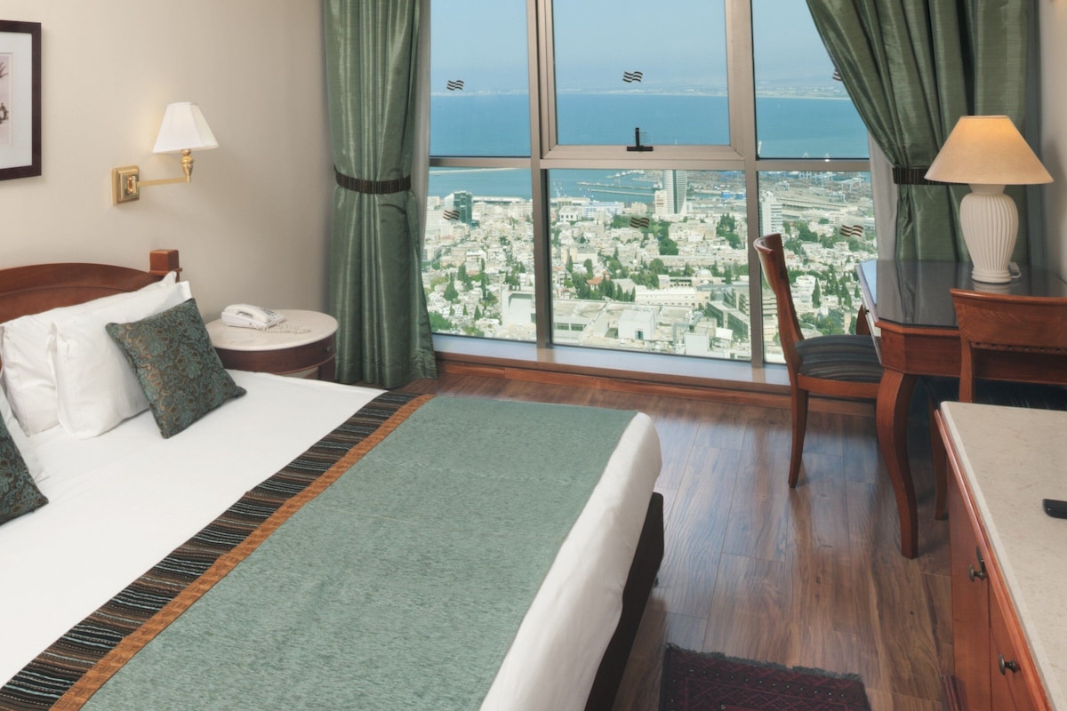 A hotel room with a large bed, a desk, a chair, a view of the city, and a window with curtains.