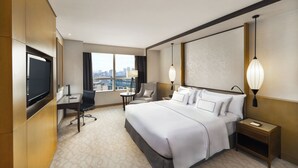 A hotel room with a bed, desk, chair, TV, bedside lamps, and a view of the city. - Melia Hanoi (Hanoi)