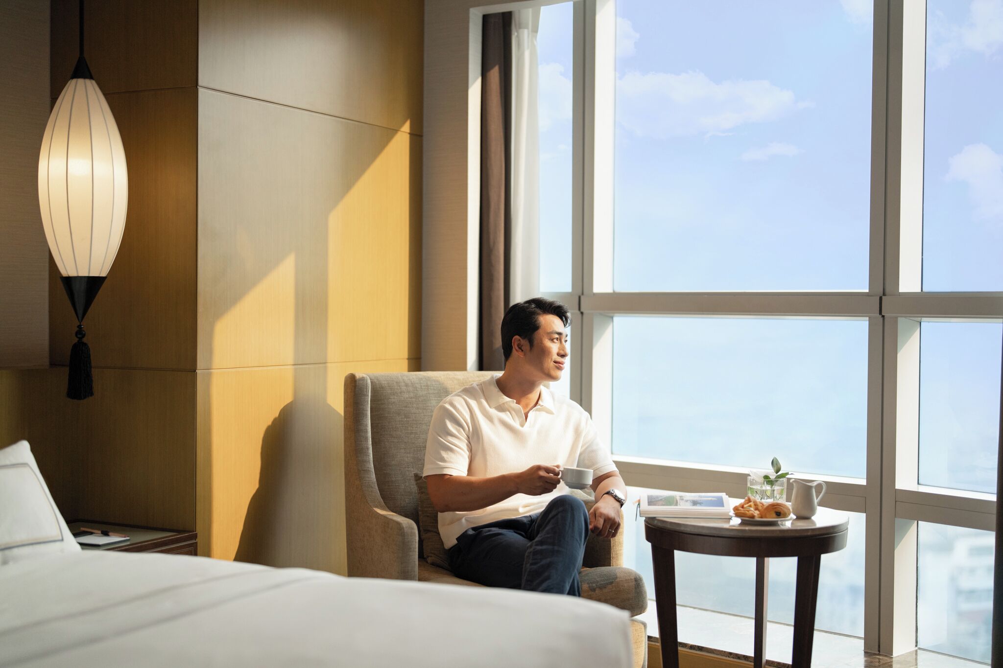 A hotel room with a large window, a bed, a chair, and a small table with a cup and a plant.