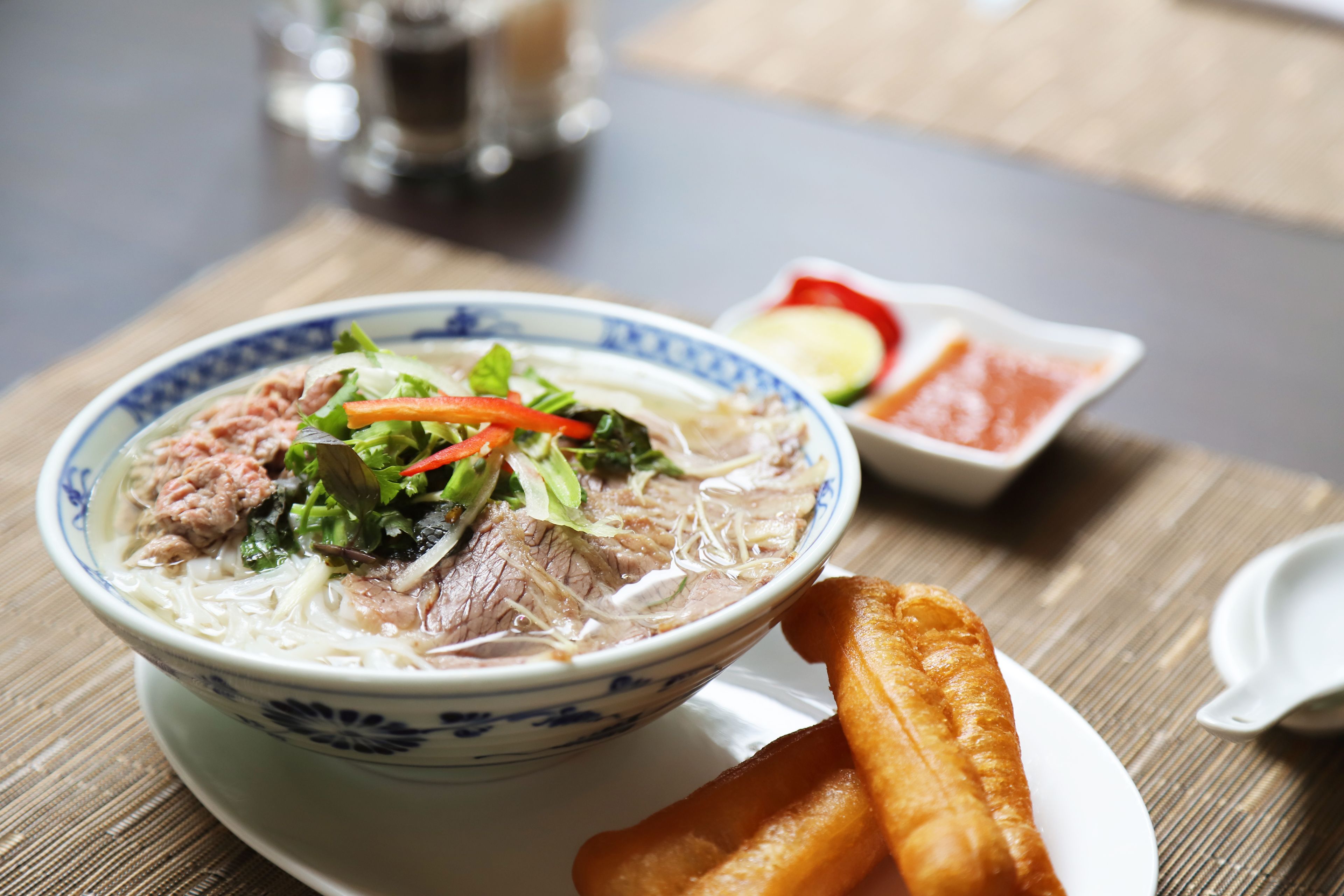 Un tazón de sopa de fideos con carne y vegetales, acompañado de una pequeña fuente de condimentos y frituras sobre una mesa de madera.