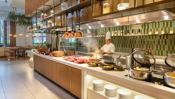 A modern kitchen with a chef preparing food, various cooking stations, and a buffet setup.