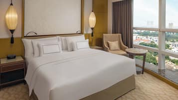 A hotel room with a large bed, two bedside lamps, a chair, a small table, and a view of the city through large windows.