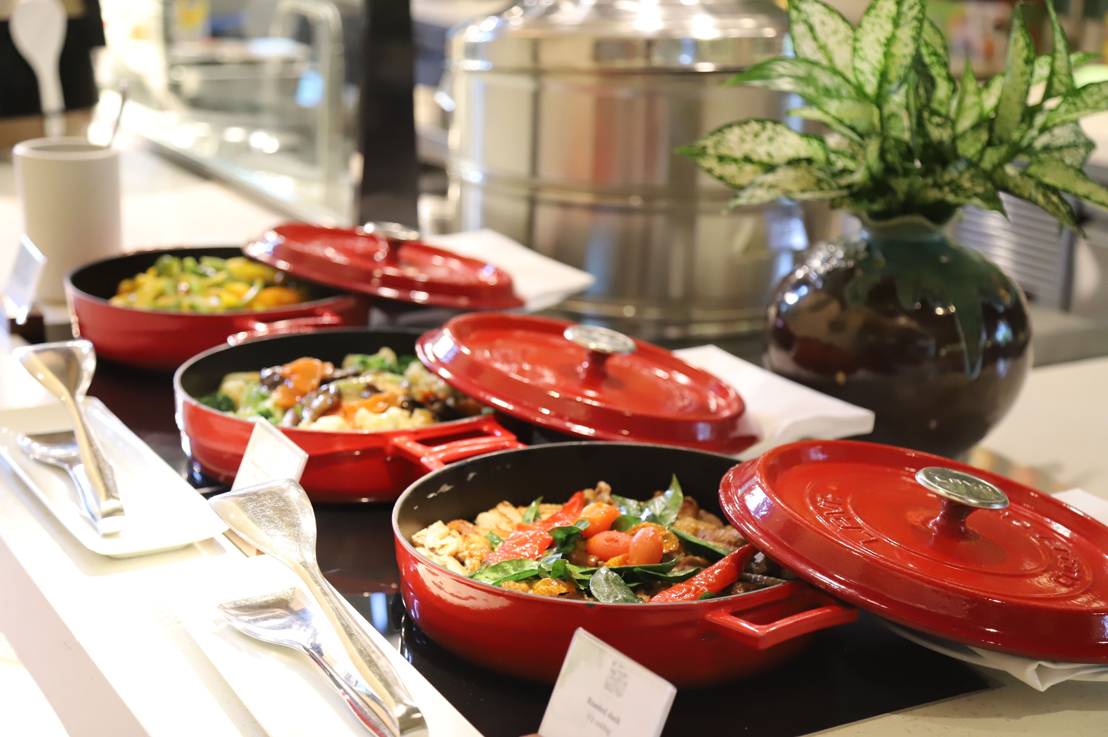 A buffet with red serving dishes containing various dishes, a large metal pot in the background, and a potted plant on the right.