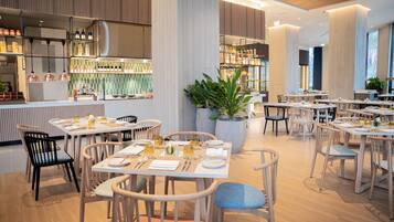 A modern restaurant interior with wooden tables and chairs, a bar area, and large potted plants.