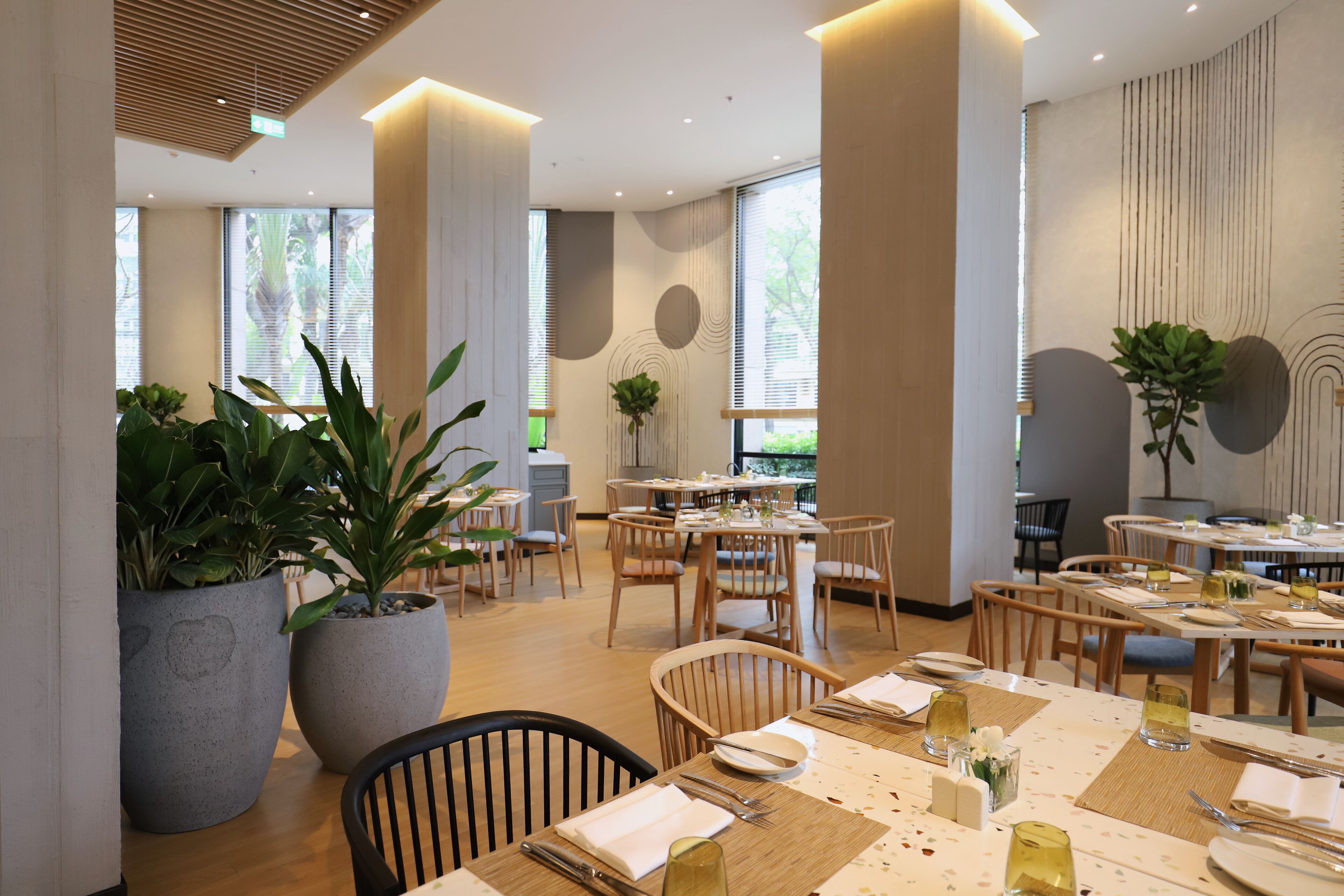 A modern dining area with wooden tables and chairs, large potted plants, and large windows allowing natural light.