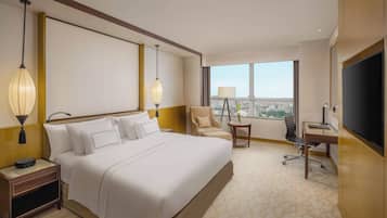 Quarto de hotel com uma cama grande, uma escrivaninha, uma cadeira, televisão e vista da cidade através de grandes janelas.