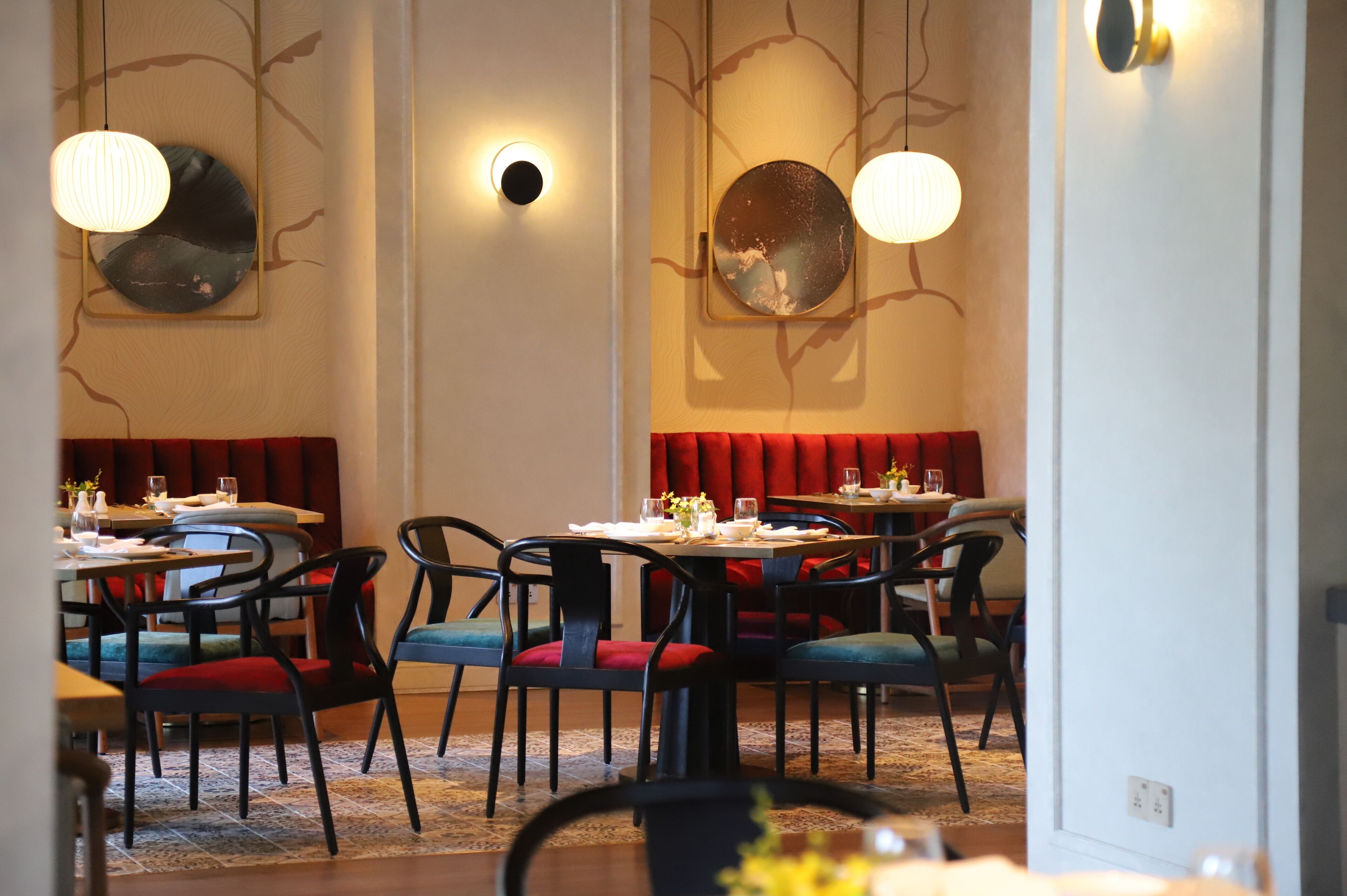 A restaurant with booths and tables, some with red cushions. There are hanging lights and a wall with a tree design.