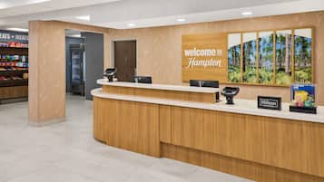 Un comptoir d’accueil d’hôtel avec une pancarte « Bienvenue à Hampton », un réfrigérateur rempli d’en-cas et une affiche présentant le programme de fidélité « Hilton Honors ».