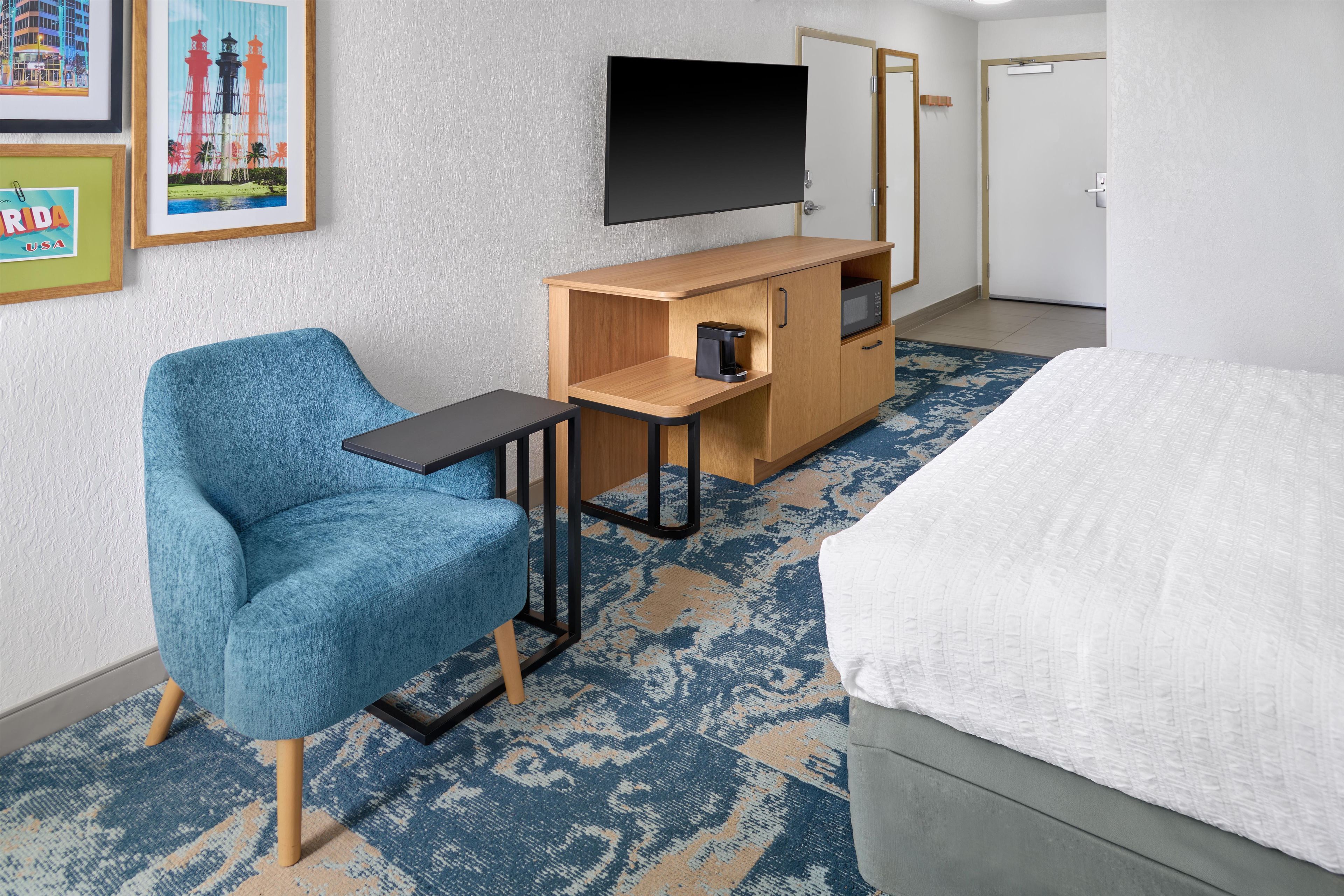 A hotel room with a bed, a desk with a coffee maker, a chair, and a TV on the wall.