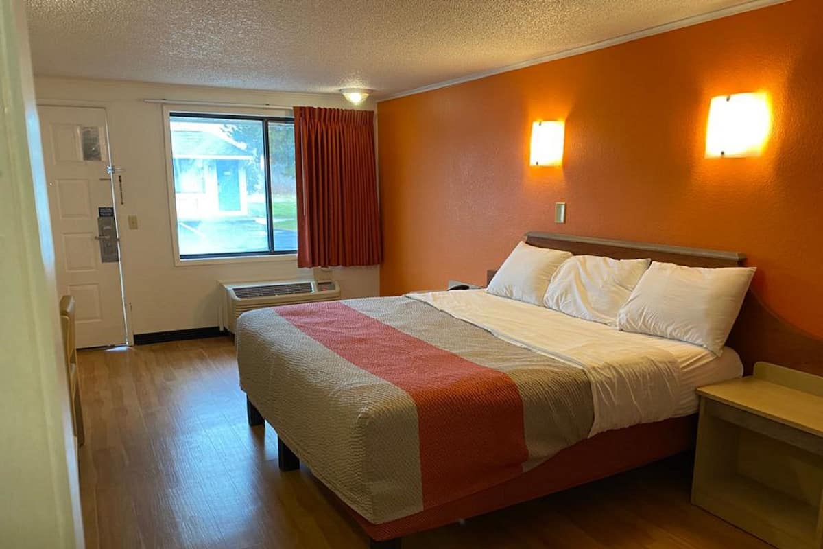 A hotel room with a bed, a bedside table, a window with curtains, and a door.