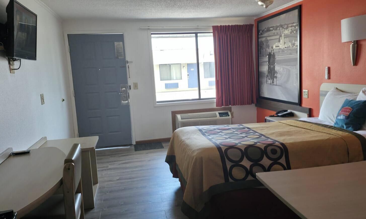 Habitación de hotel con cama, escritorio, silla, televisor, ventana con cortinas y lámpara de pared.