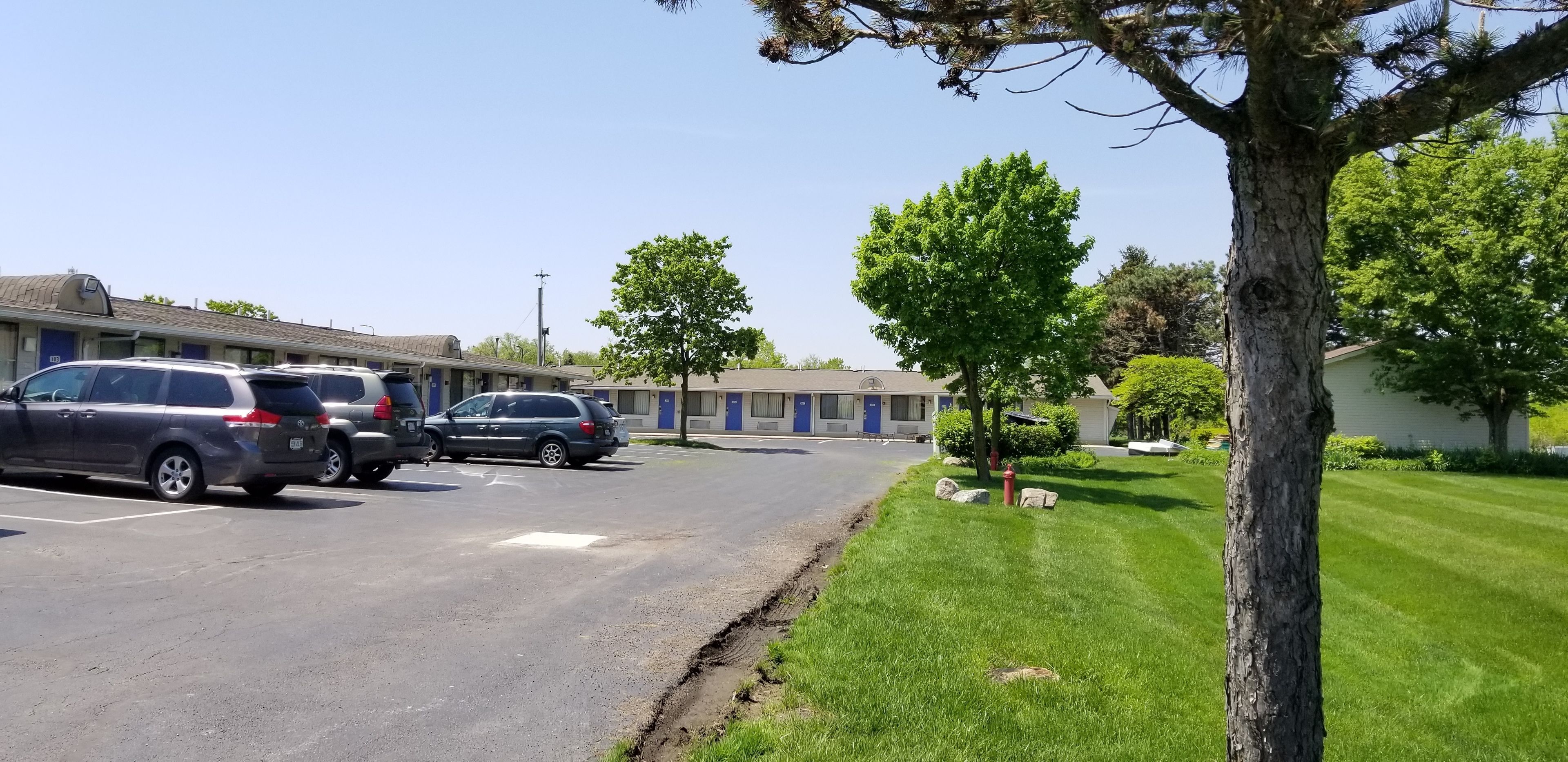 A parking lot with several cars, a building with blue doors, and a well-maintained lawn with trees.