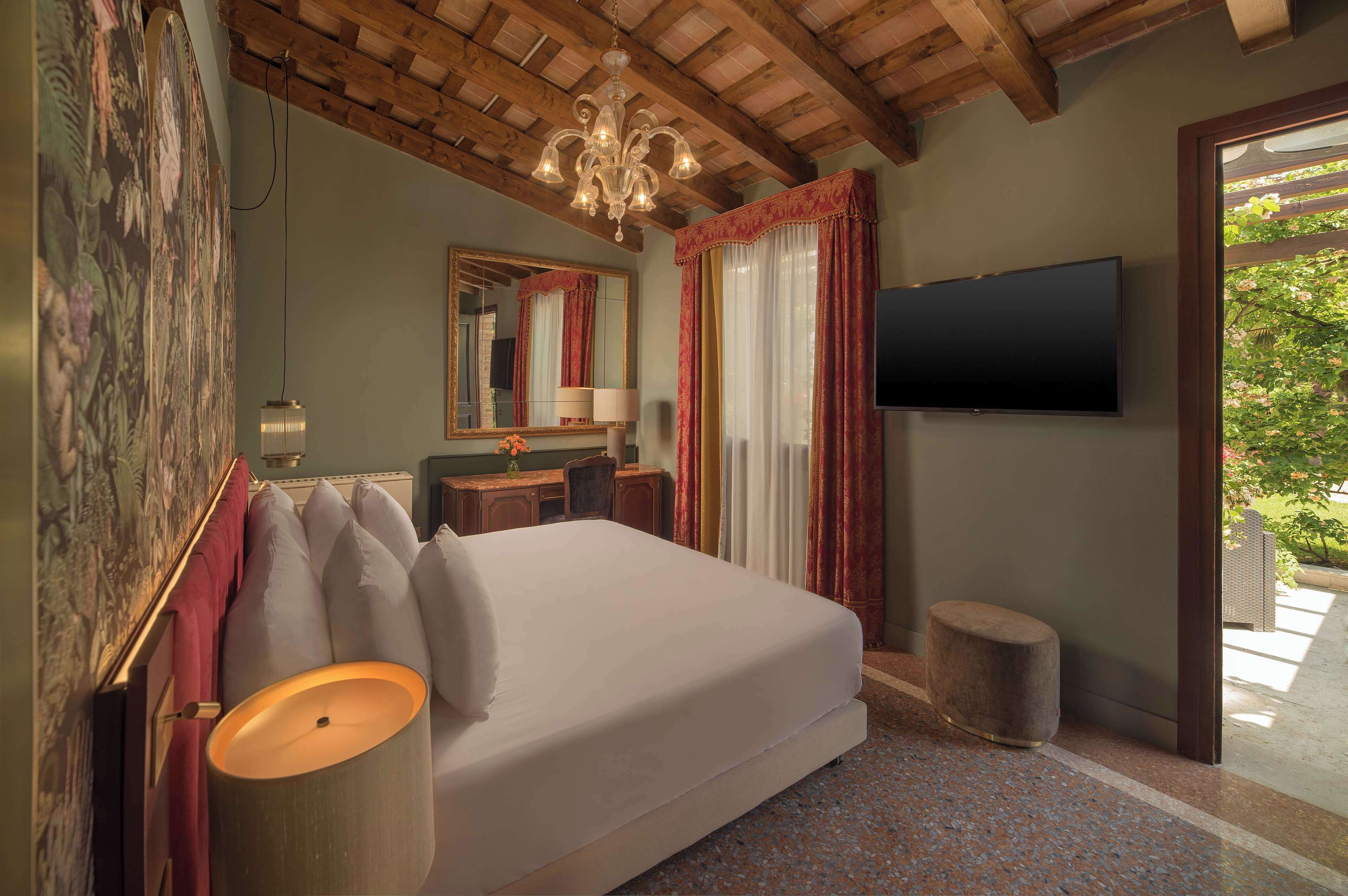 Un dormitorio con una cama grande, televisor, escritorio con lámpara y vista a zonas verdes.