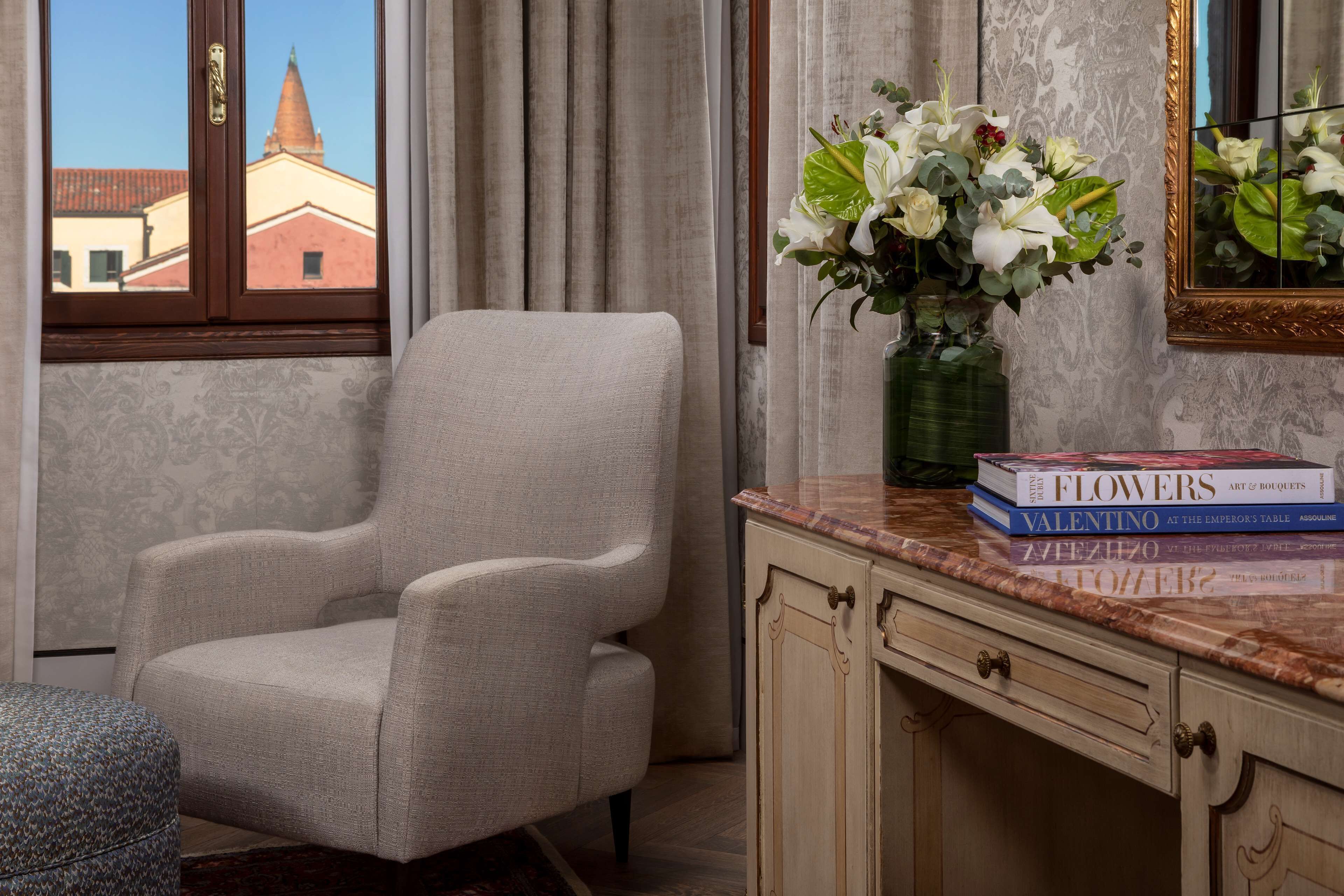 Una habitación con vistas a un edificio a través de una ventana, una silla, un florero con flores y una encimera de mármol con libros.