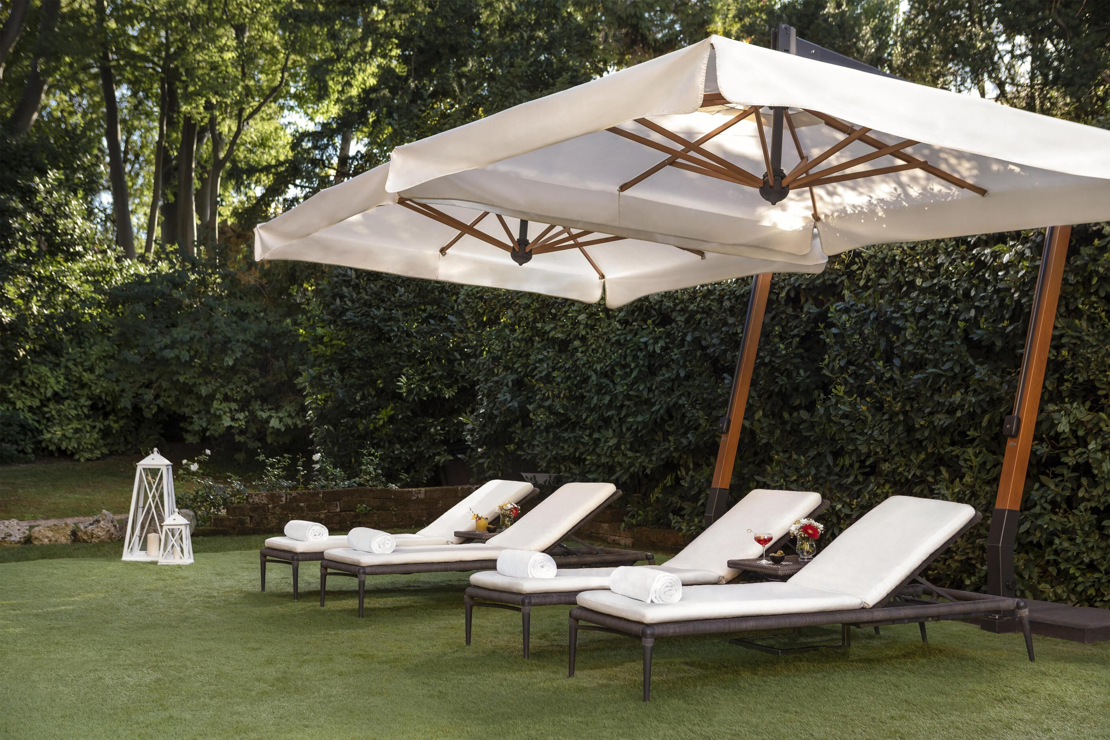 Ein Garten mit drei weißen Liegestühlen unter einem großen Sonnenschirm, umgeben von viel Grün.