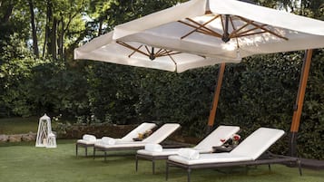Ein Garten mit drei weißen Liegestühlen unter einem großen Sonnenschirm, umgeben von viel Grün.